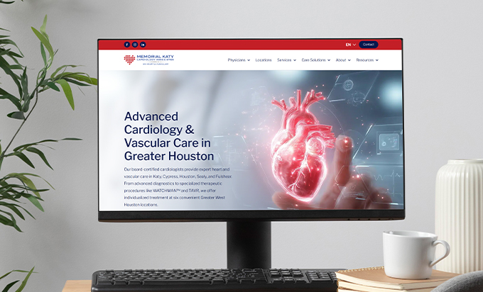 A computer monitor on a desk displays the Memorial Katy Cardiology Associates website with a glowing 3D heart graphic.